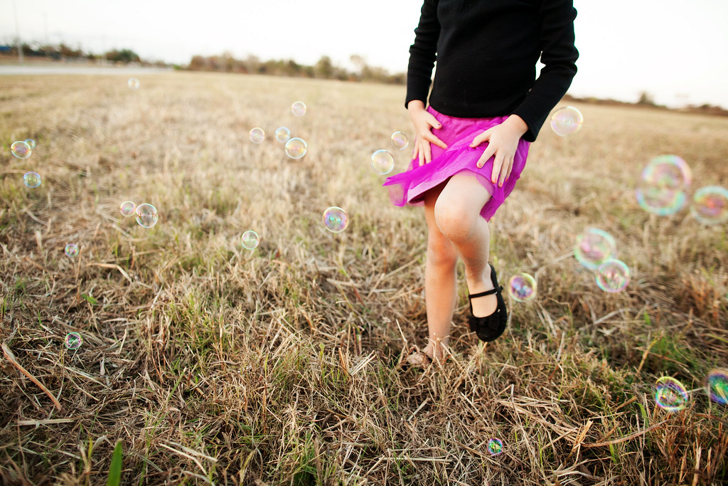 Close crop of a child's legs in black shoes and a pink skirt standing in grass, surrounded by floating bubbles in soft daylight